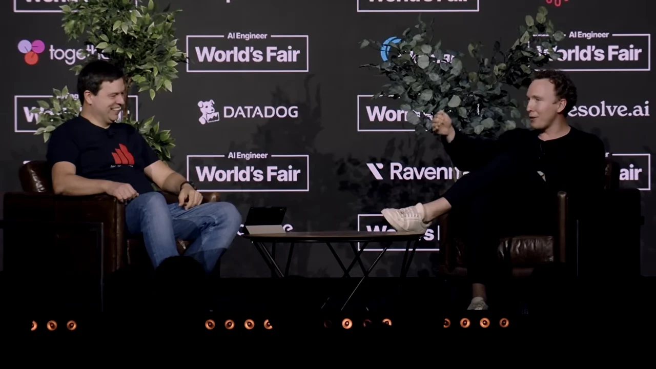 Two seated participants face each other across a low table; the participant on the left smiles while the other gestures.