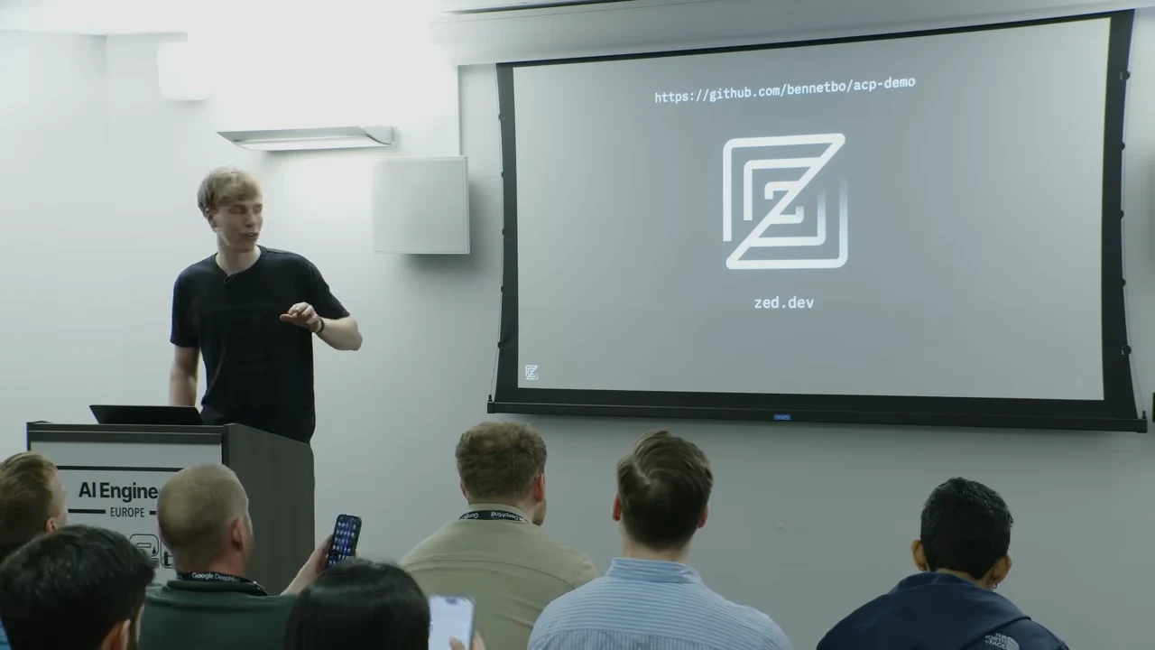 Presenter beside a projected slide showing a GitHub repository address, the Zed logo and zed.dev.
