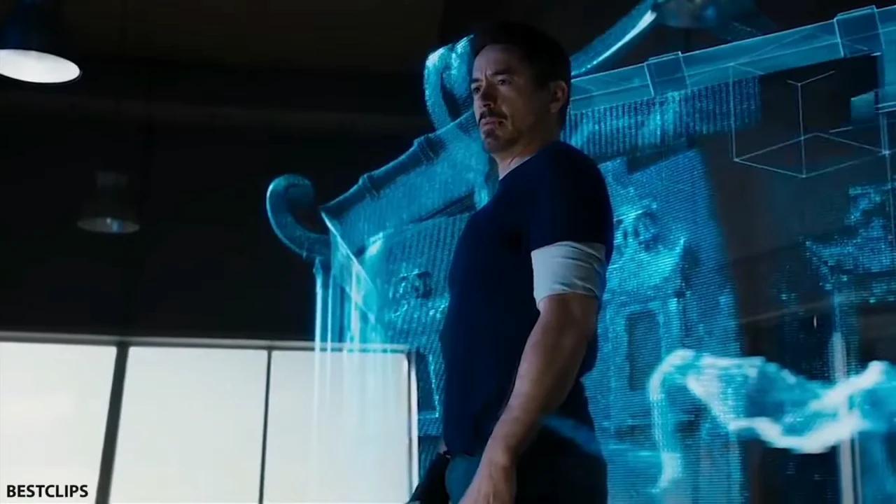 Tony Stark stands in front of a blue holographic structure in a workshop.
