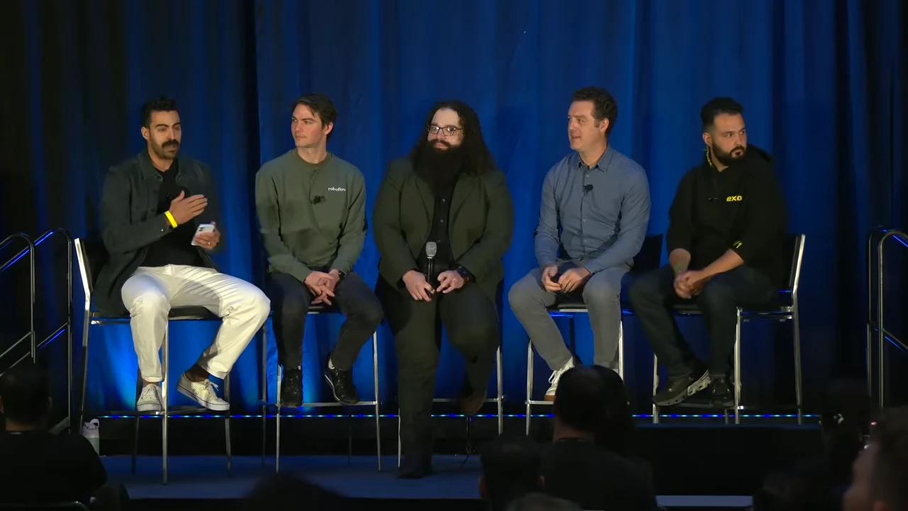 Five people sit on stools against blue curtains; the leftmost panelist gestures toward the others.