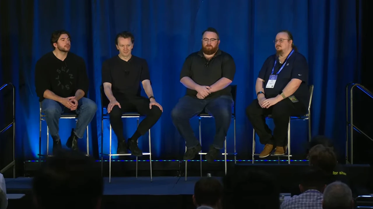 Four panelists sit on tall stools, with audience members visible along the bottom of the frame.
