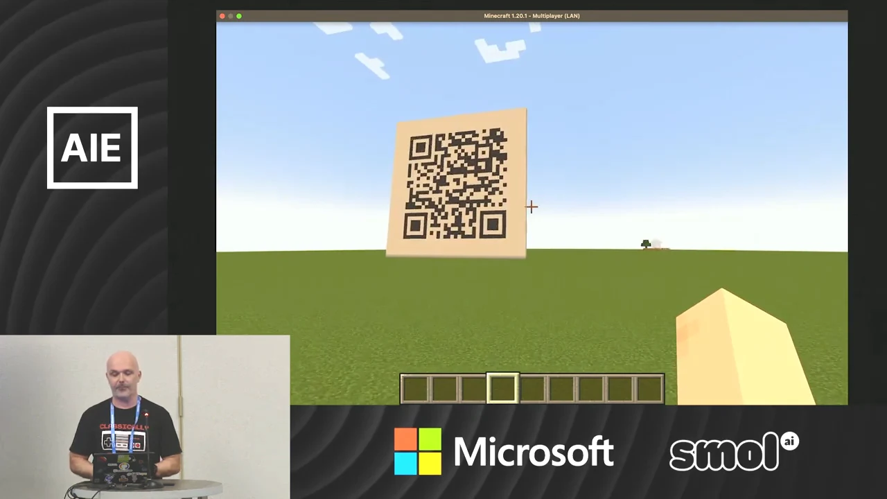 A large QR code on a pale square stands above a grassy Minecraft landscape, with the speaker inset at lower left.