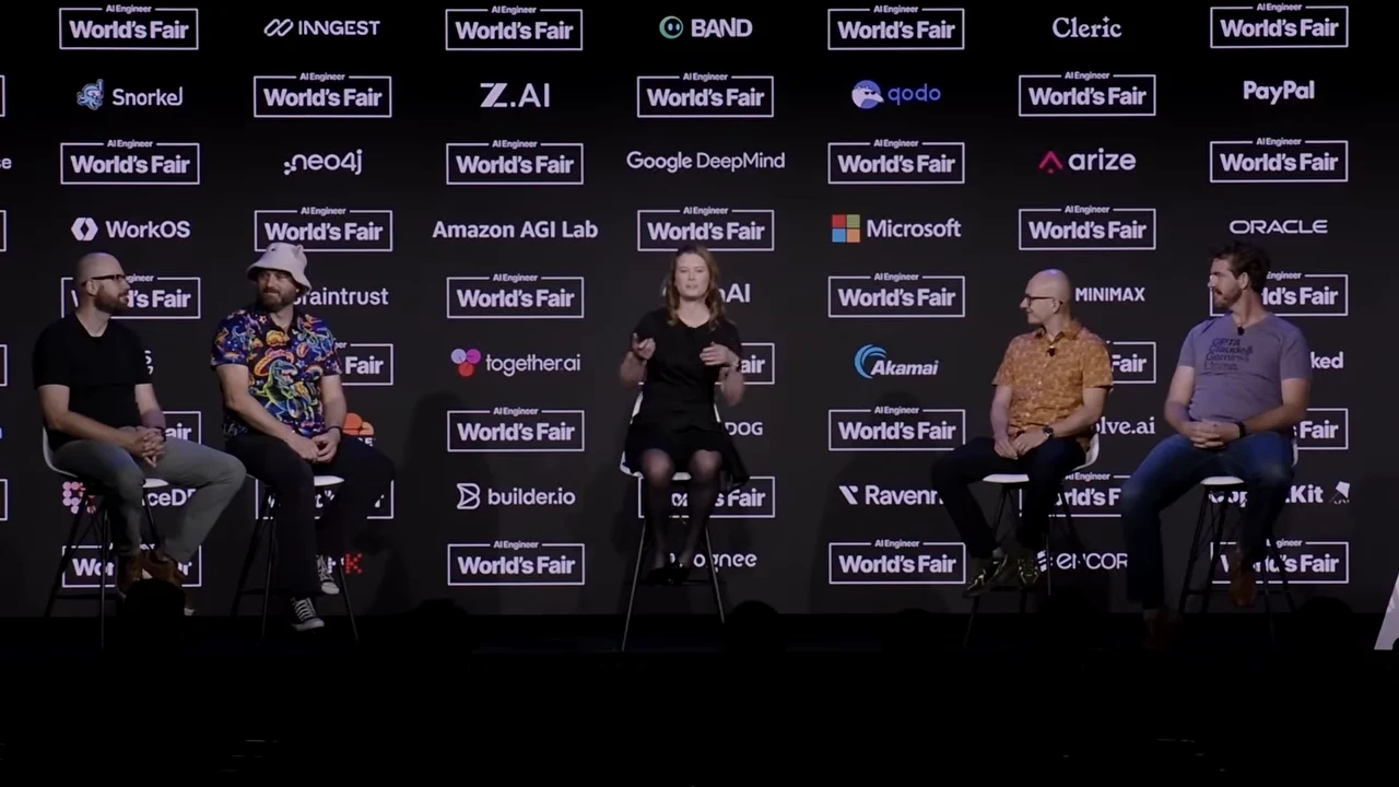 Five people seated on stools before a sponsor backdrop, with the center participant gesturing.