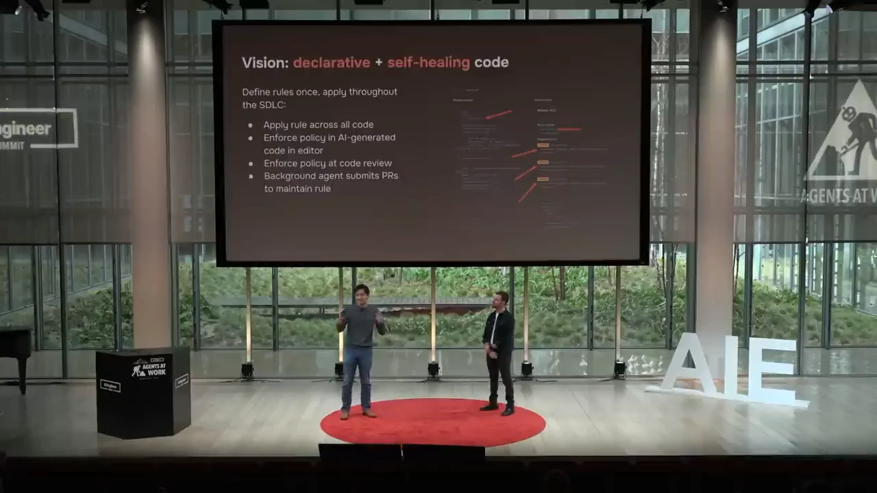 Wide stage view showing a slide about applying rules across code, enforcing policy in the editor and code review, and background agents submitting pull requests.